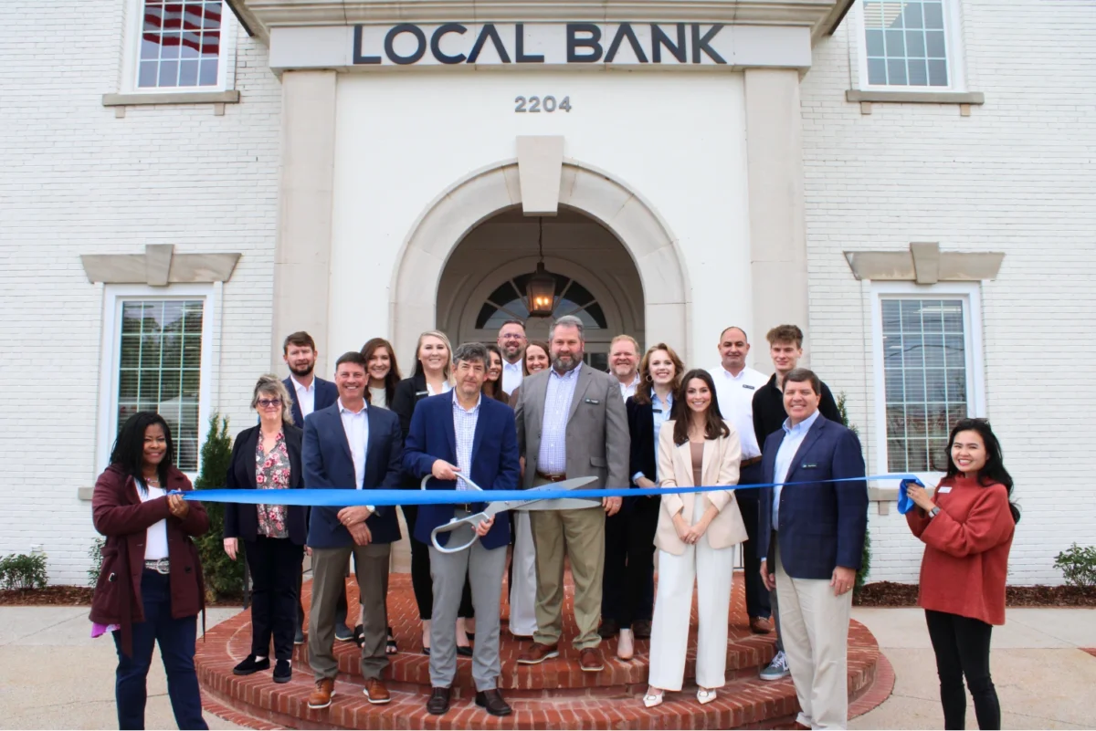 Group photo, standing in front of a bank for a ribbon cutting ceremony, about to cut a blue ribbon.